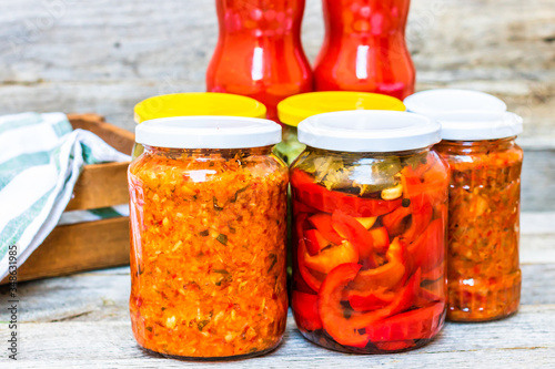 Glass jars with pickled red bell peppers and pickled cucumbers (pickles) isolated. Jars with variety of pickled vegetables, jars with zacusca and bottles with tomatoes sauce. Preserved food concept. photo