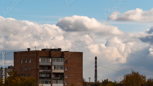 Residential building made of brick against the background of a factory chimney, clouds and blue sky on a Sunny, clear day. Houses with balconies and Windows are surrounded by trees with yellow leaves.