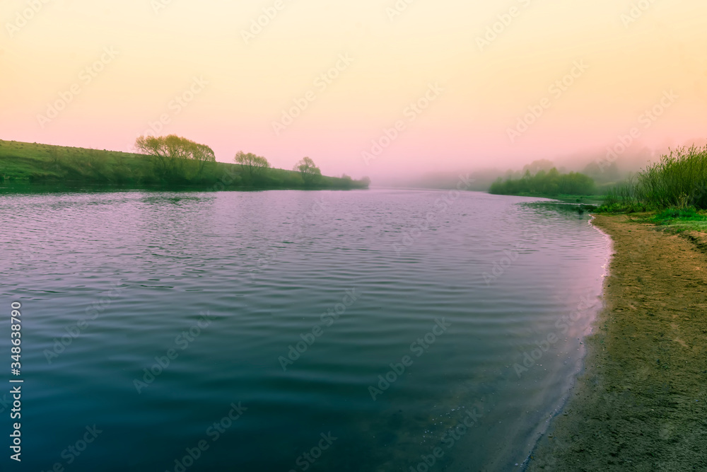 Scenic view at beautiful misty spring morning on a river beach with green grass and picturesque fog , trees and water waves, spring valley landscape