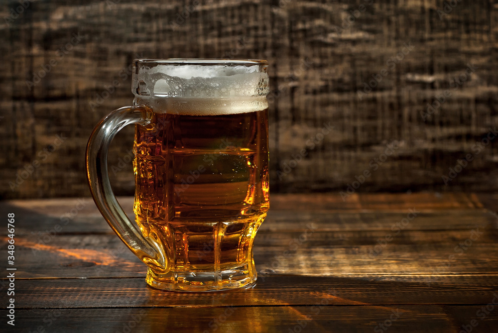 A glass of amber beer. Glasses on a wooden table. Black shabby boards on the background. Bright contrasting light and glare on the glass. Place for text near the booze. A glass of amber beer. Glasses on a wooden table. Black shabby boards on the background. Bright contrasting light and glare on the glass. Place for text near the booze.
