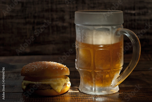 Cheeseburger with a glass of beer on a dark wooden table. Food on a black shabby background. Contrasting yellow light sparkles on the glass. Frozen mug with amber beer.