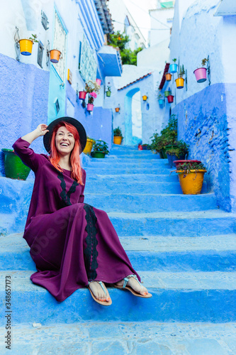 A young beautiful laughing red-haired girl in a hat and a burgundy oriental-style dress sits on the steps on a street of Chefchaouen in Morocco. © viktoriia1974