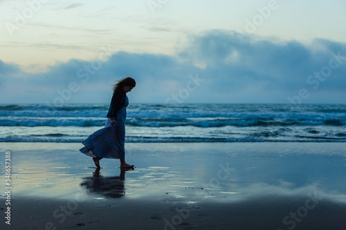 Fototapeta A sad woman with long hair in a long dress is walking at sunset along the shore of the sea or ocean