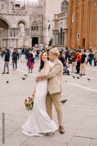 Wedding in Venice, Italy. The bride and groom are hugging in Piazza San Marco, overlooking Campanila and St. Mark Cathedral, among a crowd of tourists.