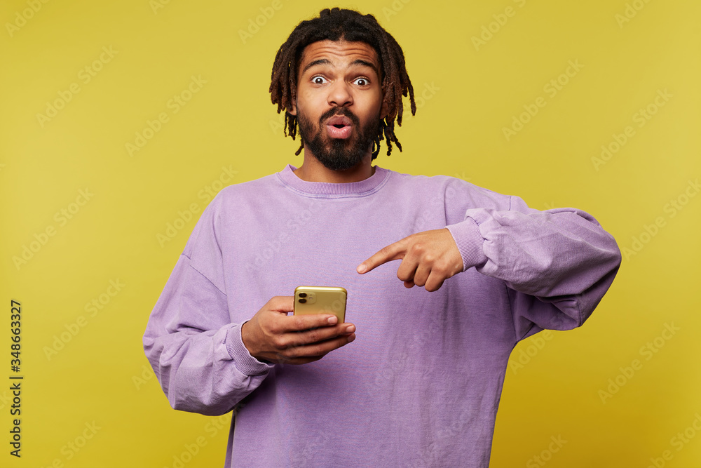 Bemused young handsome brunette male with dark skin rounding surprisedly his brown eyes and showing on his smartphone with raised forefinger, posing over yellow background