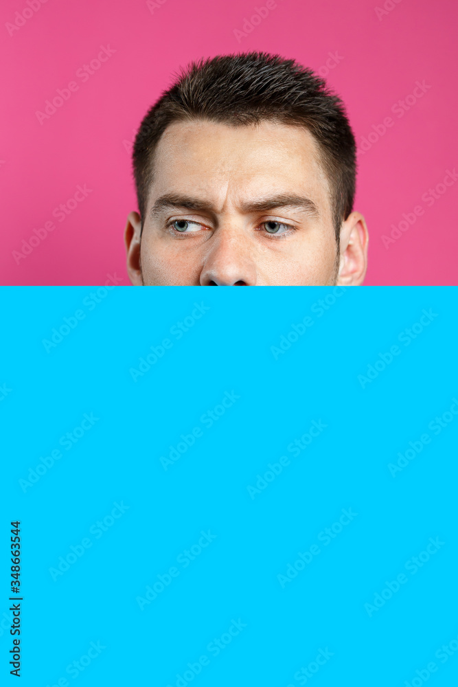 Young handsome man holding a blank cardboard blue banner over an isolated pink background, serious face, thinking about a question. Place for an inscription. Idea for recording thoughts Young handsome man holding a blank cardboard blue banner over an isolated pink background, serious face, thinking about a question. Place for an inscription. Idea for recording thoughts
