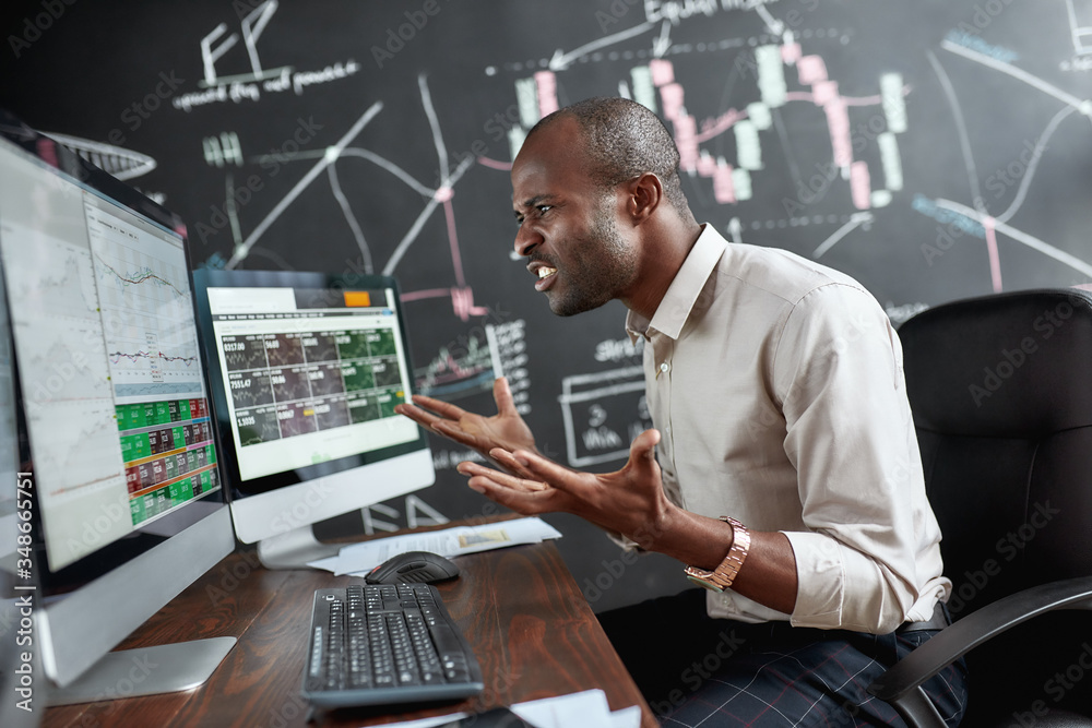 See the difference. Portrait of stylish african businessman, trader sitting by desk in front of multiple monitors. He is getting angry while looking at current graphs