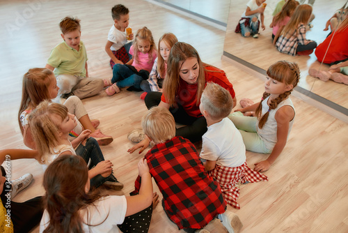 Discussing new dance perfomance. Full length portrait of young dance teacher talking to group of little girls and boys sitting on the floor in studio. Relationship between teacher and kids