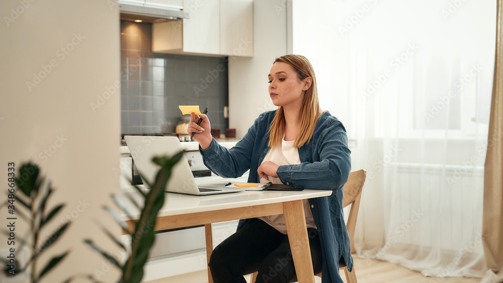 Using sticky notes. Young caucasian girl in casual clothes working from home, she is sitting at kitchen table at home and using laptop, looking at sticky note Using sticky notes. Young caucasian girl in casual clothes working from home, she is sitting at kitchen table at home and using laptop, looking at sticky note