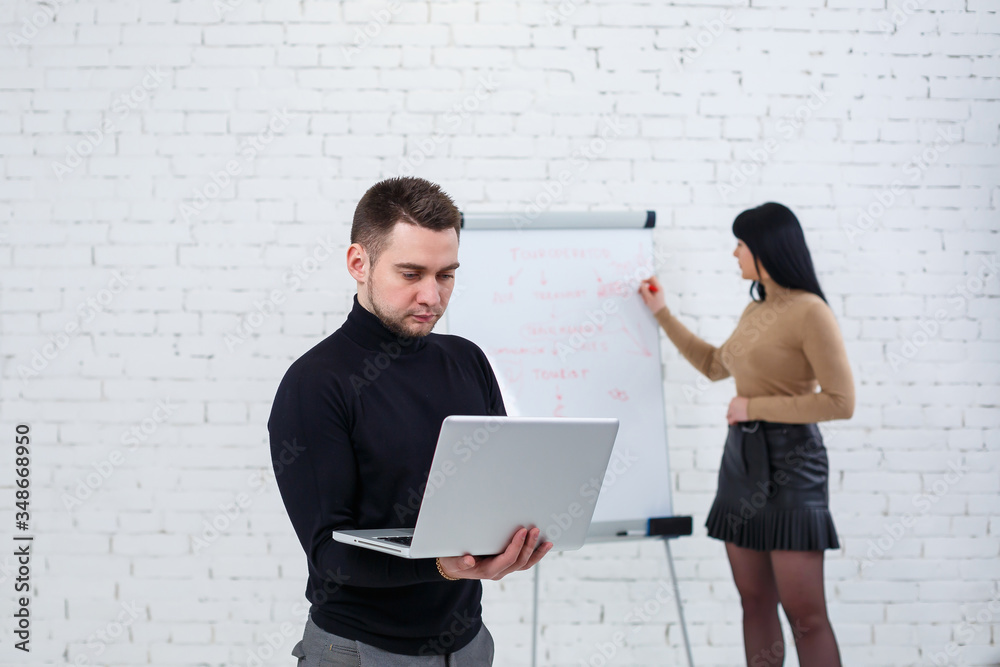 Manager businessman male announcer stands with a laptop while a woman writes a business promotion plan on a white board. A girl learns new business rules with a girl student Manager businessman male announcer stands with a laptop while a woman writes a business promotion plan on a white board. A girl learns new business rules with a girl student