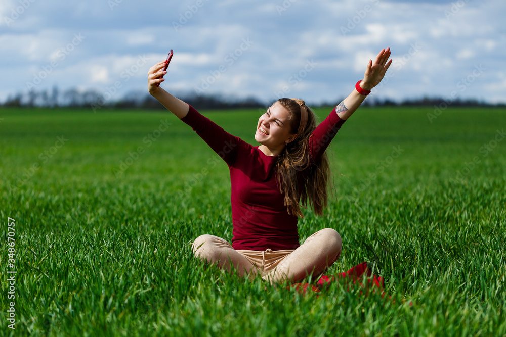 Young beautiful woman student with a phone in her hands sitting on the grass. Girl takes selfies and takes selfie pictures. She smiles and enjoys a warm day. Concept photo on smartphone Young beautiful woman student with a phone in her hands sitting on the grass. Girl takes selfies and takes selfie pictures. She smiles and enjoys a warm day. Concept photo on smartphone