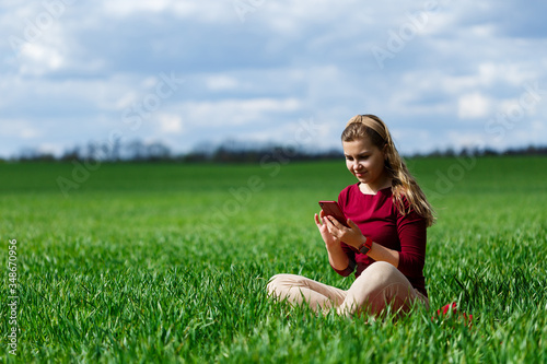 Young beautiful woman student with a phone in her hands sitting on the grass. Girl takes selfies and takes selfie pictures. She smiles and enjoys a warm day. Concept photo on smartphone © Дмитрий Ткачук