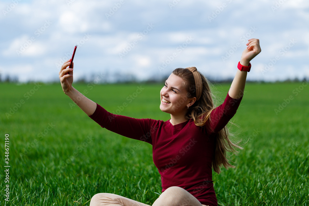 Beautiful woman with a phone in her hands sits on the grass. The girl looks at herself in the camera of the phone and takes selfie photos. She smiles and enjoys a warm day. Concept photo on smartphone Beautiful woman with a phone in her hands sits on the grass. The girl looks at herself in the camera of the phone and takes selfie photos. She smiles and enjoys a warm day. Concept photo on smartphone
