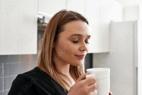 Feel good with coffee. Close up of young curvy woman closing her eyes while drinking tea or coffee in the kitchen