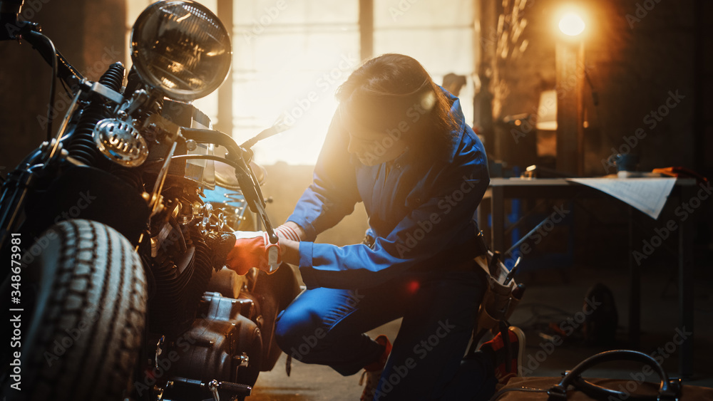 Young Beautiful Female Mechanic is Working on a Custom Bobber Motorcycle. Talented Girl Wearing a Blue Jumpsuit. She Uses a Spanner to Tighten Nut Bolts. Creative Authentic Workshop Garage.