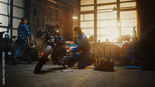 Young Beautiful Female Mechanic is Working on a Custom Bobber Motorcycle. Talented Girl Wearing a Blue Jumpsuit. She Uses a Ratchet Spanner to Tighten Nut Bolts. Creative Authentic Workshop Garage.