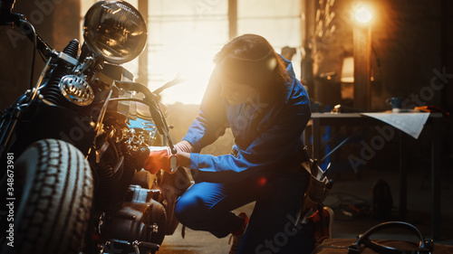 Young Beautiful Female Mechanic is Working on a Custom Bobber Motorcycle. Talented Girl Wearing a Blue Jumpsuit. She Uses a Spanner to Tighten Nut Bolts. Creative Authentic Workshop Garage.