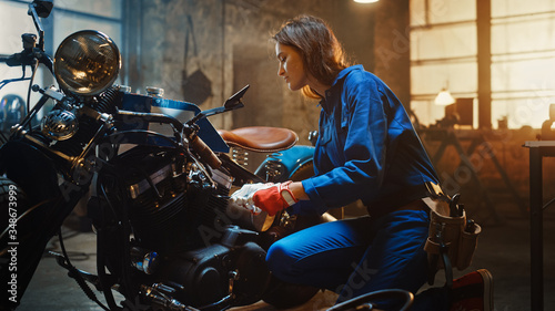 Young Beautiful Female Mechanic Comes Working on a Custom Motorcycle in Garage. Talented Girl Wearing a Blue Jumpsuit. She Uses a Ratchet to Tighten Nut Bolts. Creative Authentic Workshop.