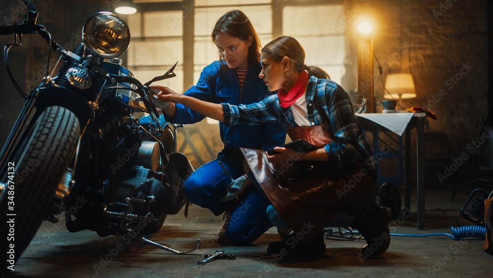 Two Young Beautiful Female are Discussing The Work Done on a Custom Bobber Motorcycle. Talented Girls Use a Tablet Computer. They are In Good Spirit and Happy. Creative Authentic Workshop Garage.