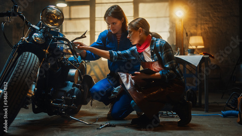 Two Young Beautiful Female are Discussing The Work Done on a Custom Bobber Motorcycle. Talented Girls Use a Tablet Computer. They are In Good Spirit and Happy. Creative Authentic Workshop Garage.