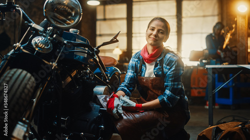 Young Beautiful Female Mechanic is Working on a Custom Bobber Motorcycle. Talented Girl Wearing a Checkered Shirt and an Apron. She Smiles at the Camera. Creative Authentic Workshop Garage.