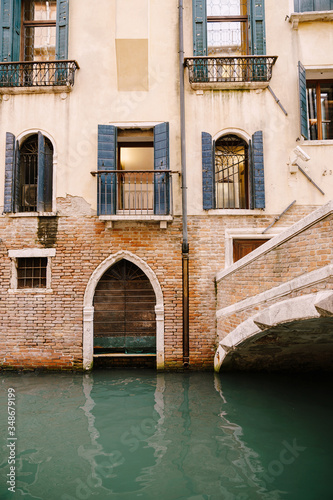 A brick building in the water in Italy, Venice. A brick bridge over a small narrow canal, classic Venetian windows in the facade of the building with wooden shutters and forged bars.