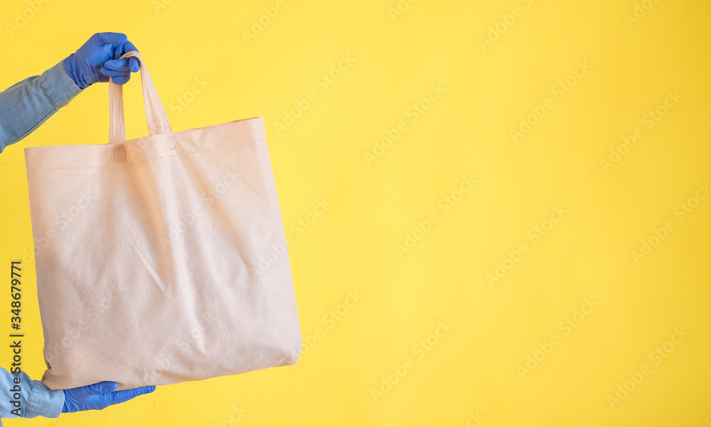 Sending eco-friendly reusable cotton bag on a yellow background. The courier holds a canvas fabric bag without plastic packaging on a yellow background. Safe delivery to the epidemic. Sending eco-friendly reusable cotton bag on a yellow background. The courier holds a canvas fabric bag without plastic packaging on a yellow background. Safe delivery to the epidemic.