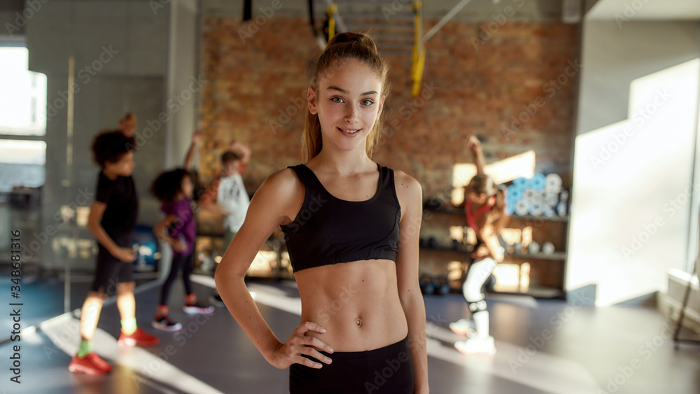 Sportive and Beautiful. Portrait of a girl smiling at camera before warming up, exercising together with other kids and trainer in gym. Sport, healthy lifestyle, active childhood concept