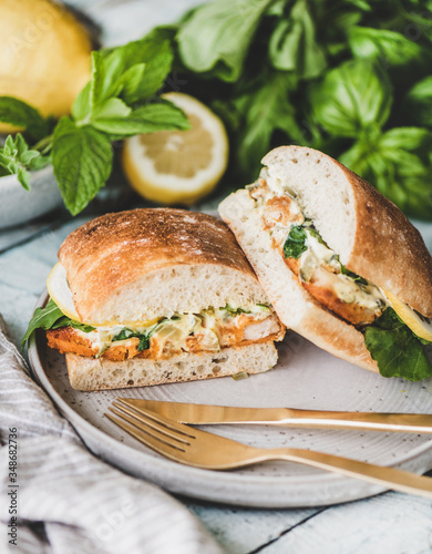 Breakfast table with fresh fried fish sandwich with tartare sauce, lemon and arugula cut in halves on plate, fresh greens, close-up. Healthy easy breakfast ideas