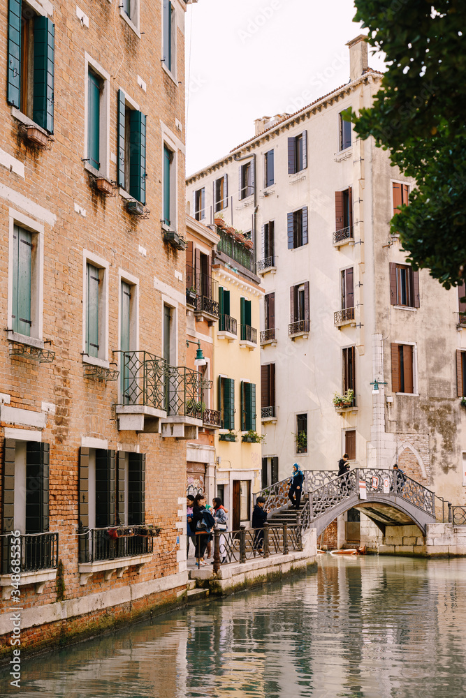 Tourists walk on an arable bridge over a small Venetian canal, against the backdrop of apartment buildings on the streets of Venice, Italy.