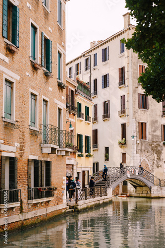 Tourists walk on an arable bridge over a small Venetian canal, against the backdrop of apartment buildings on the streets of Venice, Italy.