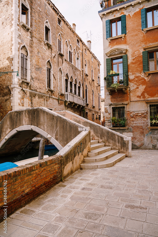 A beautiful stone bridge with a staircase across the Venetian Canal on the streets of Venice, in Italy. The facades of beautiful brick buildings with Venetian-style windows and wooden shutters.