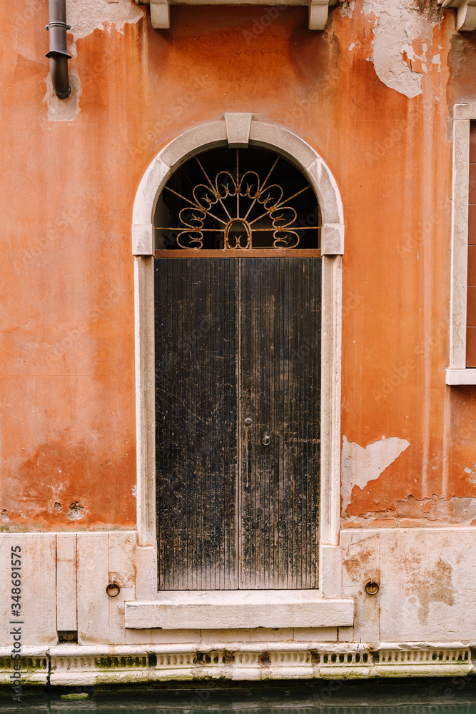 Close-ups of building facades in Venice, Italy. An old black, scratched door, in an arched doorway, with a rusty forged grille at the top, in an orange building