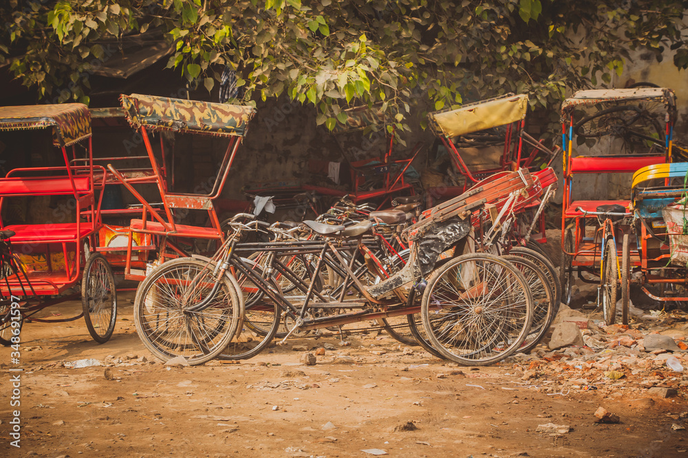 Parked bicycle rickshaws in india, multitude of rickshaws in red color and bicycles waiting to be used on a gravel parking lot.