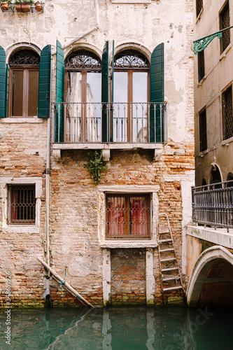 Close-ups of building facades in Venice, Italy. Beautiful Venetian windows with open wooden green shutters. An old red brick house next to a bridge over a small narrow canal.