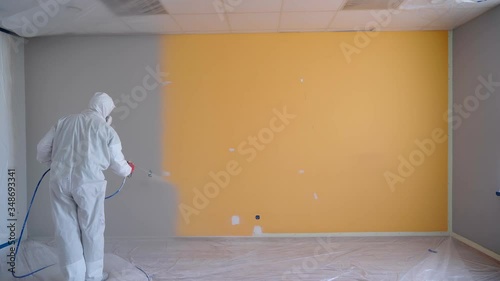 Worker wearing white workwear painting the orange wall in white color by airless spray gun. Airless Spray Painting. Repair at home or office. photo