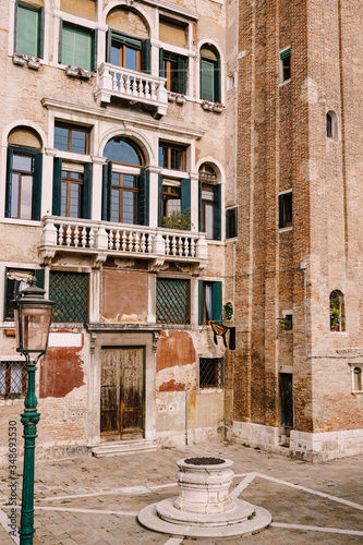 Close-ups of building facades in Venice, Italy. An old street well in the square in front of a brick house. There are many Venetian-style windows on facade of building. Vintage street lamp is green.