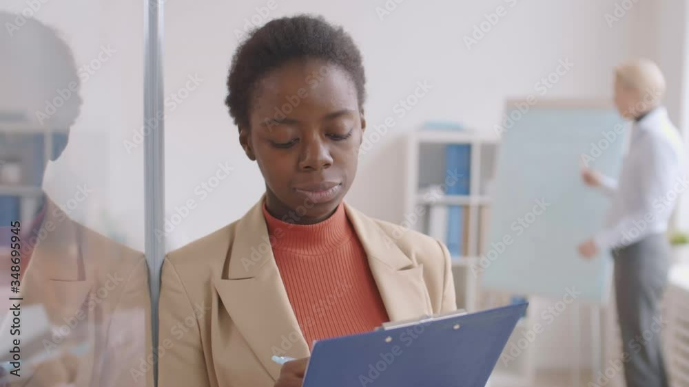Medium shot portrait of young African woman standing in office with clipboard in her hands and looking at camera while her colleague working on background Medium shot portrait of young African woman standing in office with clipboard in her hands and looking at camera while her colleague working on background