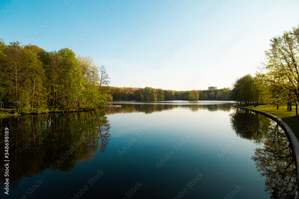 A lake in the city park early in the morning. Quiet and peaceful, in harmony, no people. Fresh green trees and grass, blue sky and clouds reflecting in the water. Warm sun light. Minsk Belarus