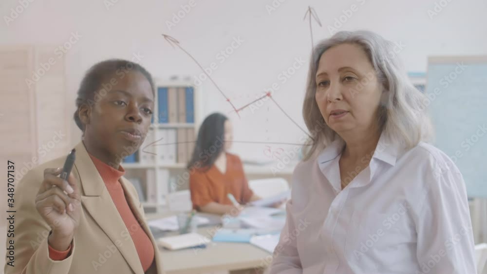 Medium shot of grey-haired Asian woman and young African woman standing together at glass wall and drawing chart on it using black markers while their colleagues working on background Medium shot of grey-haired Asian woman and young African woman standing together at glass wall and drawing chart on it using black markers while their colleagues working on background
