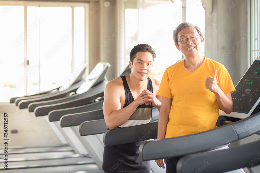 An elderly Asian man, Thai, and his trainer are standing with their thumbs at the treadmill, with a backdrop as exercise equipment in the exercise room and a copy space An elderly Asian man, Thai, and his trainer are standing with their thumbs at the treadmill, with a backdrop as exercise equipment in the exercise room and a copy space