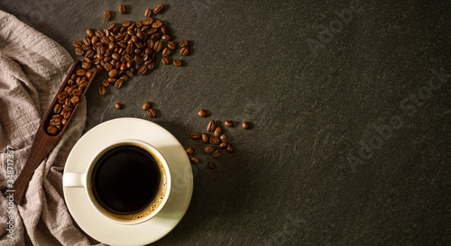 Top view above of Black hot coffee for morning with milk foam for morning menu in white ceramic cup with coffee beans roasted and apron on dark stone table background. Flat lay with copy space.