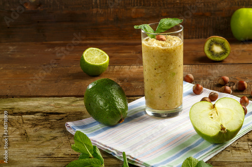 A glass of green smoothie on a wooden table on a napkin next to avocado apple lime nuts.