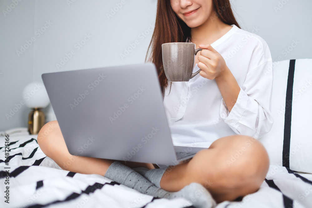 Closeup image of a beautiful woman using and working on laptop computer , drinking coffee while sitting on a white cozy bed at home Closeup image of a beautiful woman using and working on laptop computer , drinking coffee while sitting on a white cozy bed at home
