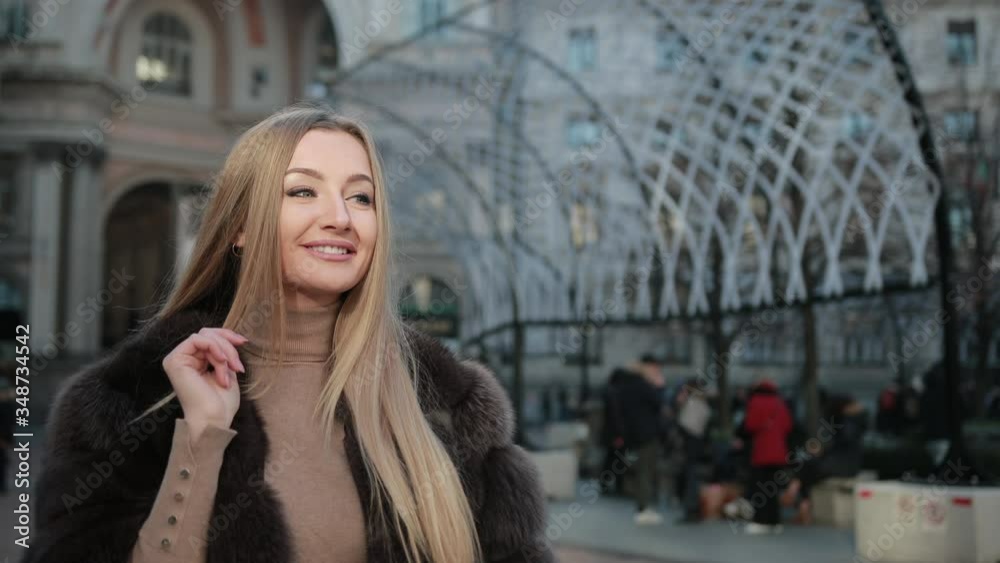 A Young Blonde Woman Is Flirting On City Street. Blowing Kisses, Waving, Smiling, Wearing Fur Coat. Bright Sunny Day. Slow Motion A Young Blonde Woman Is Flirting On City Street. Blowing Kisses, Waving, Smiling, Wearing Fur Coat. Bright Sunny Day. Slow Motion