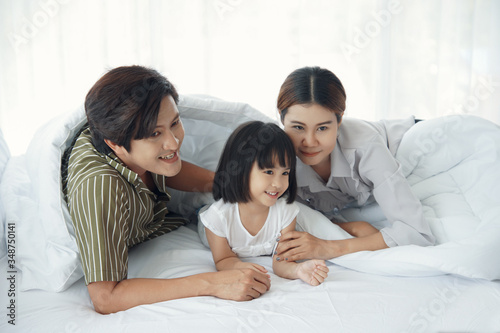 Happy asian young family spend time together. Dad mom daughter are smiling lying under white blanket on the bed at bedroom
