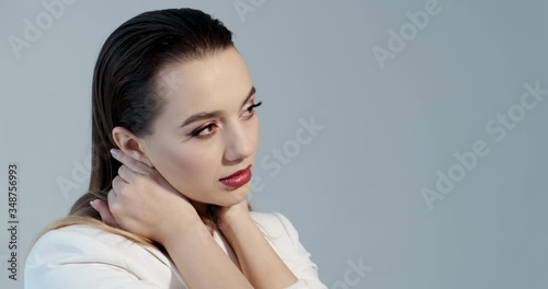 Slow motion of posing model in photo studio, close-up, girl smooths her hair with hands, opens eyes, turns head, enjoys, in white jacket, burgundy lipstick on her lips, professional makeup on her face