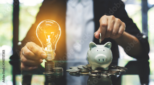 A businesswoman putting coin into piggy bank and a light bulb over coins stack on the table for saving money and financial concept photo