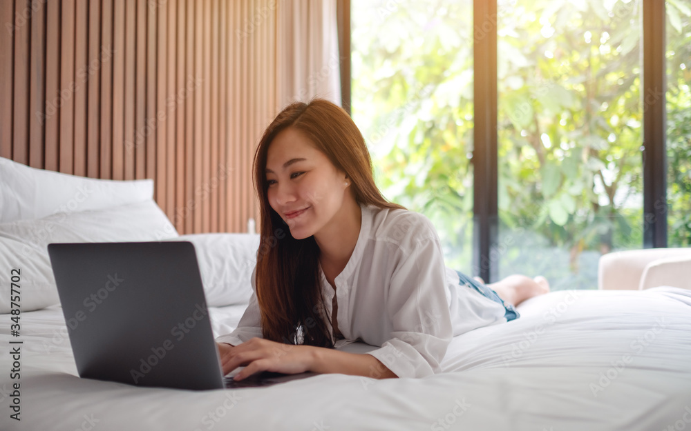 A beautiful asian woman using and working on laptop computer while lying down on a white cozy bed at home A beautiful asian woman using and working on laptop computer while lying down on a white cozy bed at home