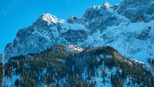 The mountainous Alpine landscape. High snow-capped mountains with sharp peaks, rocks and cliffs under a clear blue sky. Large coniferous forest at the foot of the mountains. Winter season of skiing. 